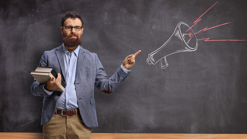 professor com livros na mão em frente a quadro de ardósia numa sala de aula